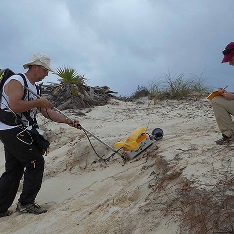 A man using a yellow georadar machine on a beach.