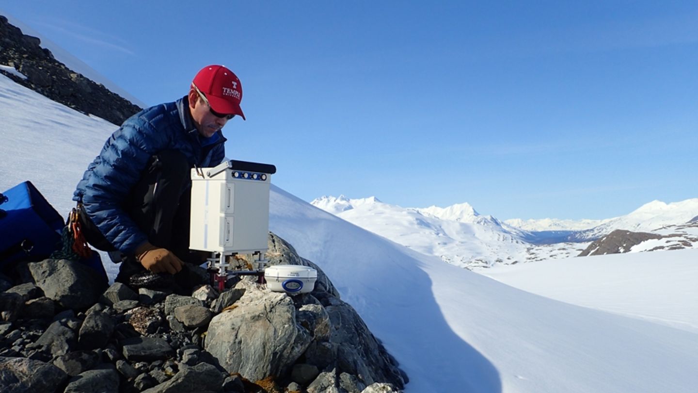 Temple University Assistant Professor Atsuhiro Muto at work on another research project earlier this year, examining changes of the Wolverine Glacier in Alaska. A man in a red Temple baseball cap perched on rocks with a snowy backdrop.